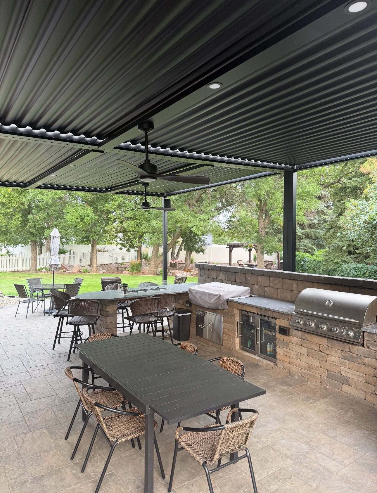 Black louvered pergola over a paver patio with an outdoor kitchen, bar seating and ceiling fans
