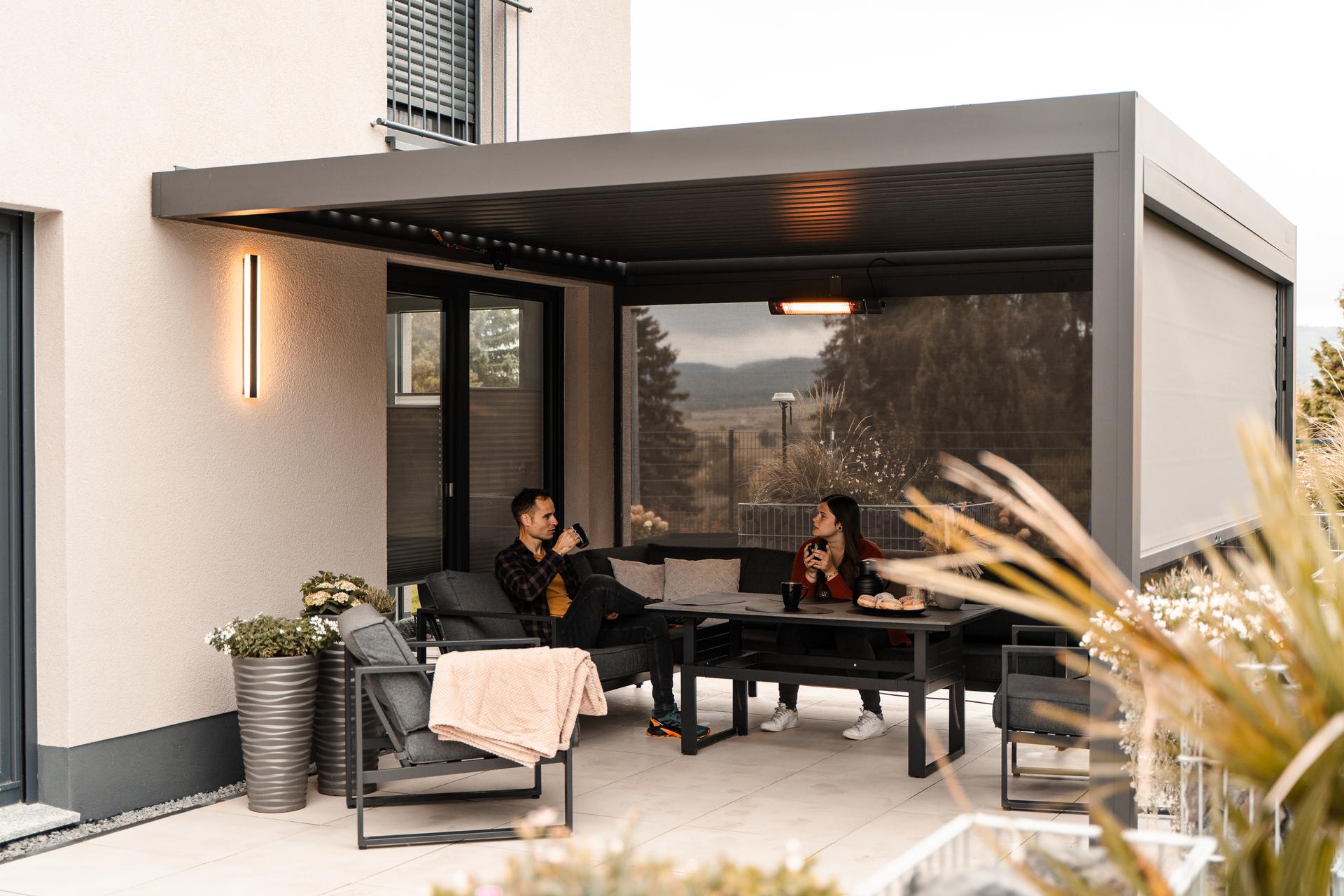 A couple under a lit louvered pergola in the evening with the overhead heater on